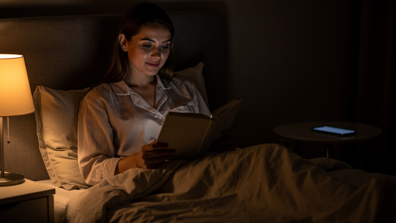 Une femme lit un livre dans une chambre sombre et apaisante avant le coucher, éclairée par une lampe de chevet chaude, tandis qu’un téléphone est posé à distance.
